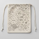 A drawstring pouch made of light beige canvas fabric, printed with a dark gray fantasy map design featuring text like 'Crimson Dawn' and 'Cursed Forest.' The pouch is laid flat on a brown wooden surface.