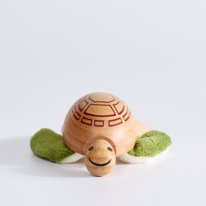 A wooden turtle toy with a natural wood shell and head, green plush flippers, and a smiling face, photographed on a clean white background.