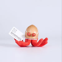 Red plush toy with a wooden egg on a white background, featuring the brand 'anomalies'.