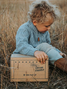 A toddler with curly light brown hair sits outdoors in dry grass, wearing a light blue cable-knit sweater and matching knit pants, leaning on a vintage-style cardboard box printed with 'LIVING FOSSILS'.
